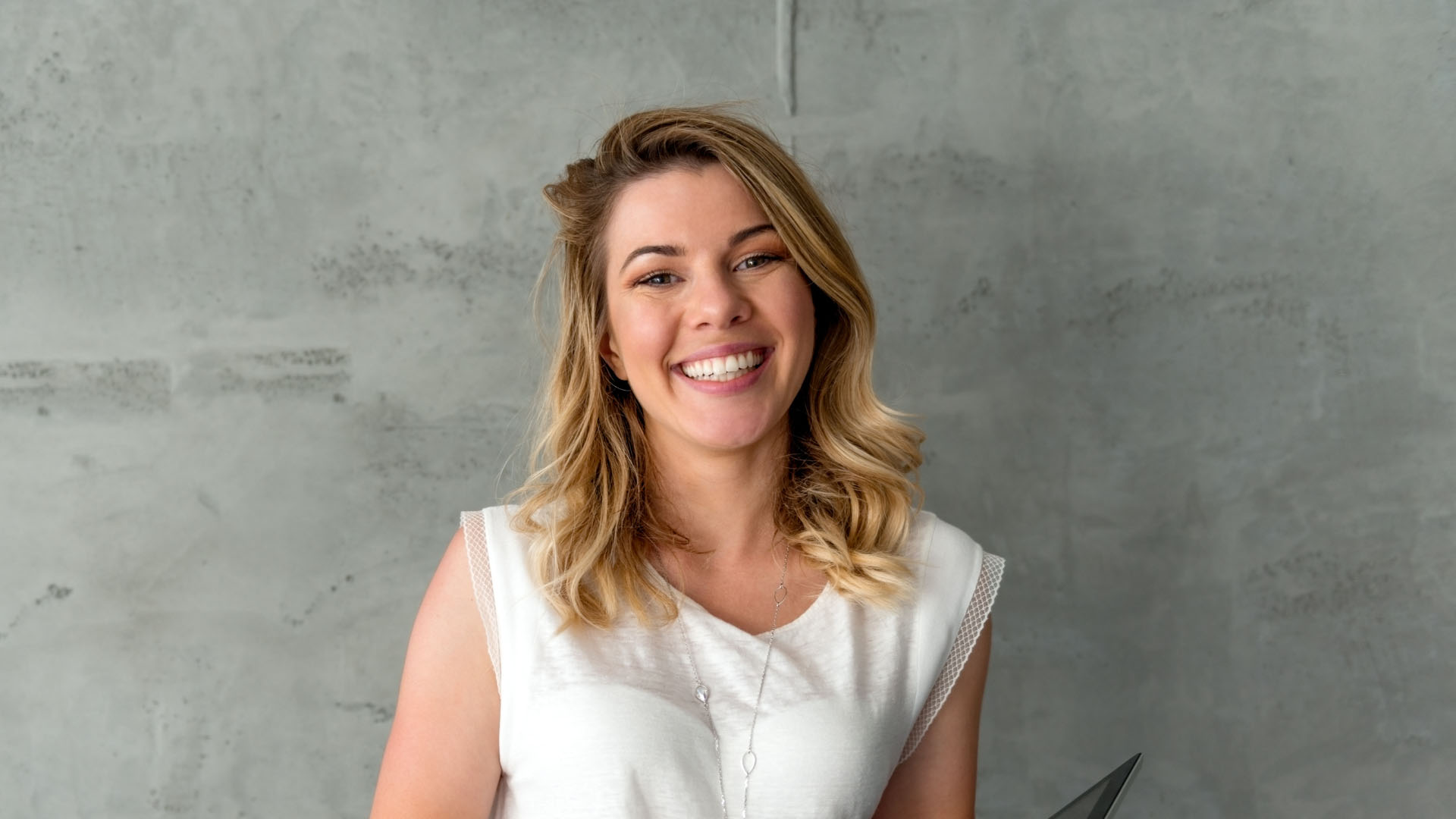 Smiling woman with blonde wavy hair wearing a white sleeveless top and silver necklace against a grey concrete wall